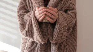 Person wearing a taupe chunky knit cardigan with cable stitches while holding a ceramic mug