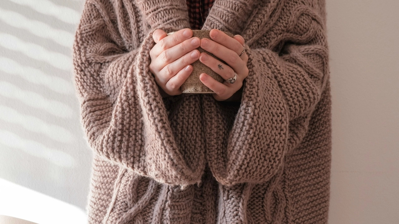 Person wearing a taupe chunky knit cardigan with cable stitches while holding a ceramic mug