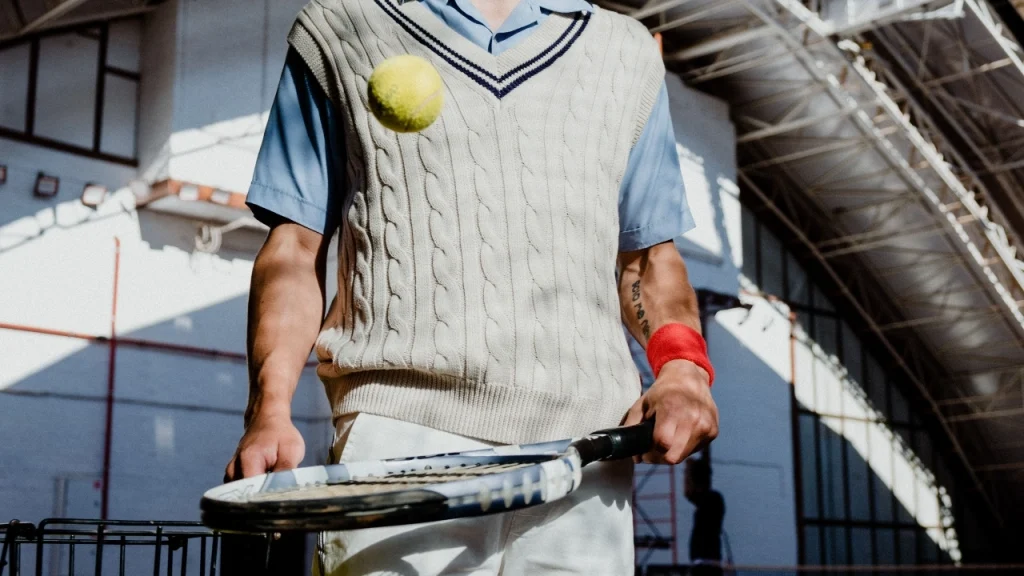 Person wearing cream custom vest knitwear with cable stitches and striped V-neck trim while playing tennis