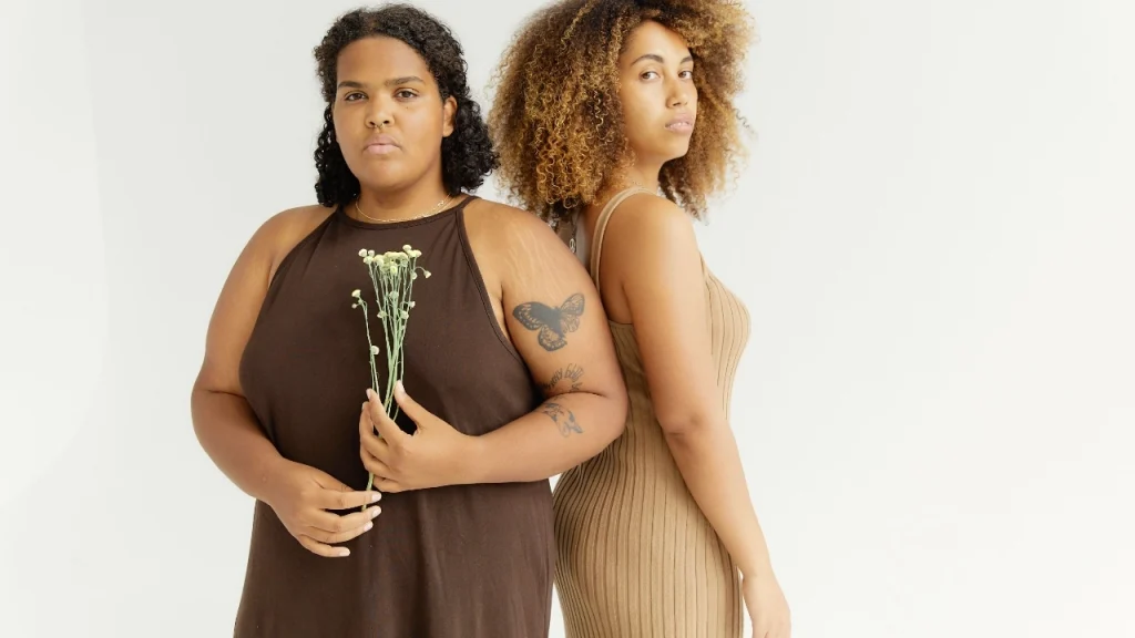 Two models wearing brown and beige knit midi dresses standing in a bright minimalist studio.