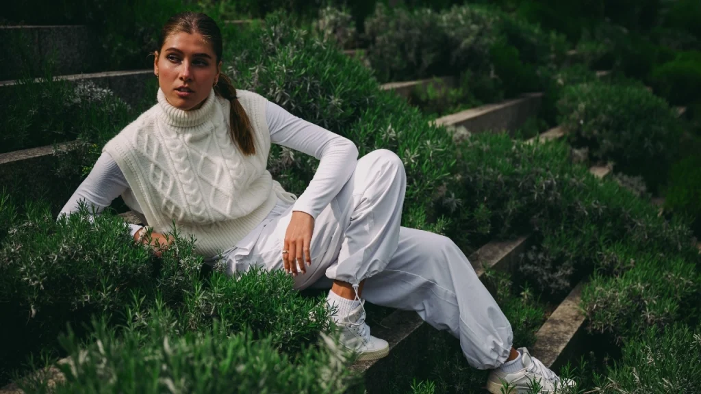 Woman wearing a white cable knitted sweater vest layered over a long sleeve top in a garden setting.