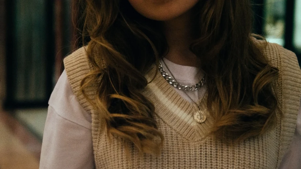 Close-up of a beige V-neck knitted sweater vest layered over a white T-shirt with silver accessories.