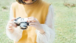 Woman wearing a yellow knitted sweater vest over a white blouse while holding a camera outdoors.