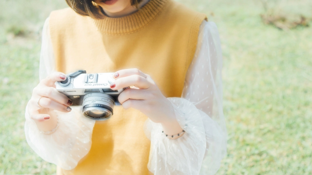 Woman wearing a yellow knitted sweater vest over a white blouse while holding a camera outdoors.