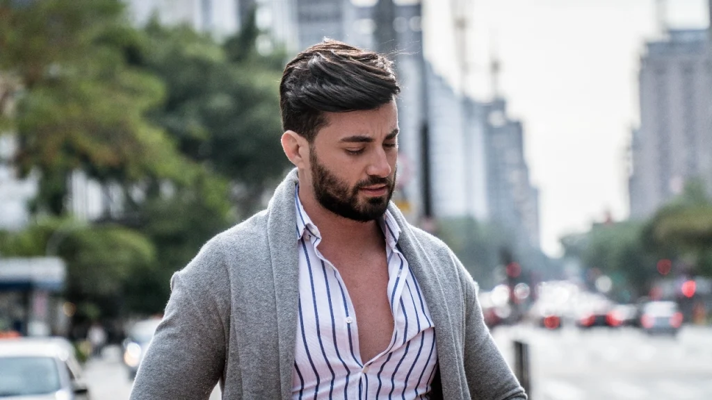 Man wearing a grey mens cardigan over a striped shirt while walking in the city.