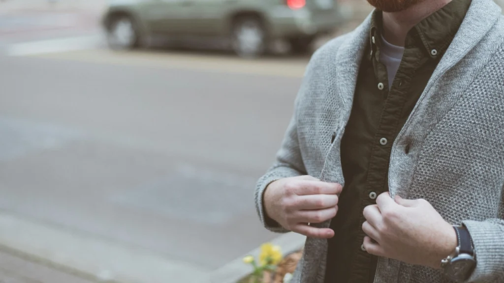 Close-up of a mens cardigan in grey knit fabric layered over a dark shirt in an urban street setting.