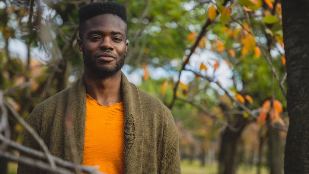 Man wearing an olive mens cardigan over an orange shirt in an outdoor park setting.