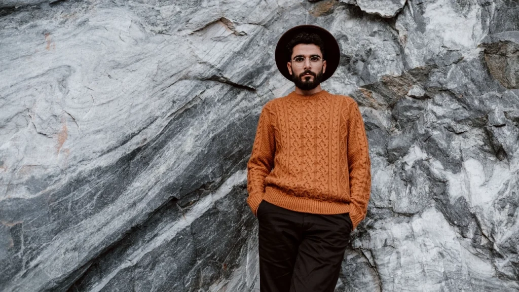 Man wearing an orange cable knit sweater against a stone wall, showing textured Mens Knitwear styling.