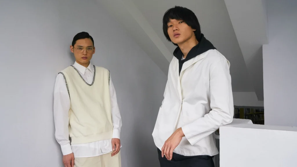 Male model wearing a white mens sleeveless jumper layered over a shirt in a minimalist studio setting.