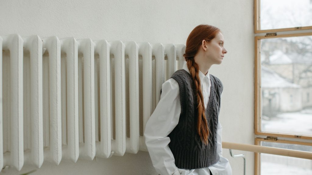 Woman wearing a dark cable knit sleeveless jumper over a white shirt beside a winter window