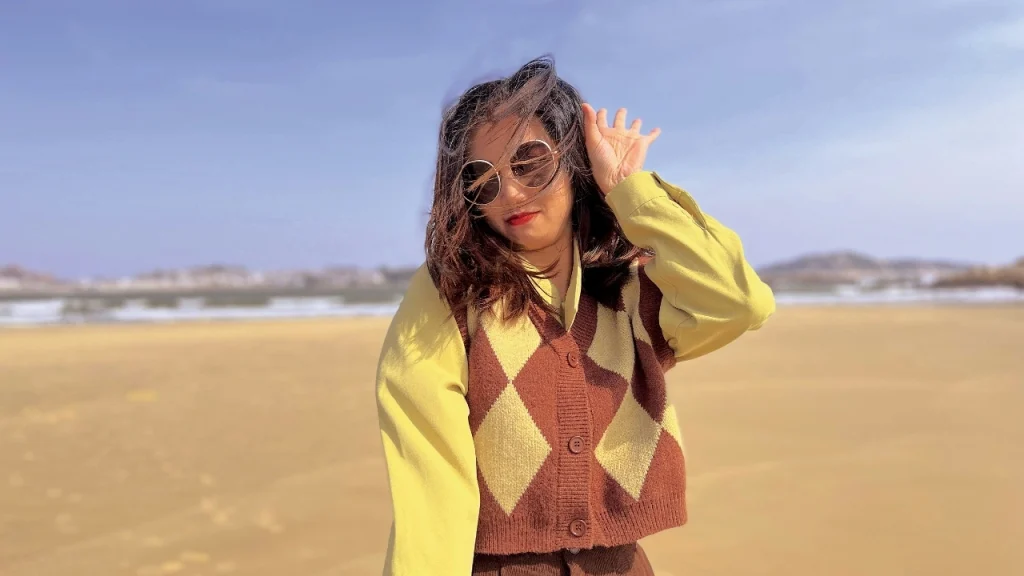 Woman wearing an argyle sweater vest over a yellow long sleeve top in an outdoor beach setting