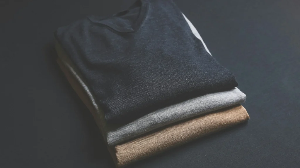 Three folded sweaters in dark, grey, and camel tones arranged on a dark surface.
