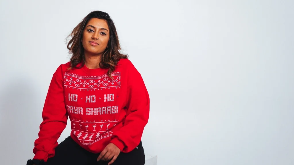 Model wearing a red custom sweater with white holiday lettering and decorative knit-style graphics in a studio setting.