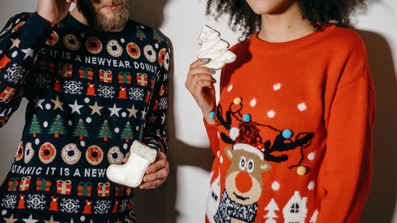 Two people wearing colorful custom sweaters with Christmas graphics while holding festive cookies.