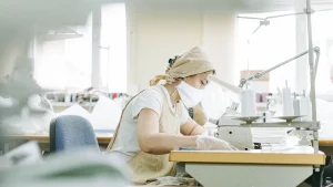 Worker wearing protective gear sewing garments in a bright sweater factory workshop.