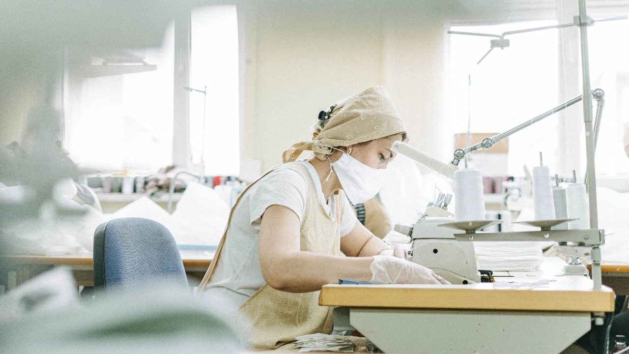 Worker wearing protective gear sewing garments in a bright sweater factory workshop.