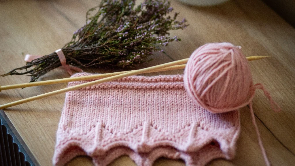 Pink Custom Knitwear swatch with yarn ball, knitting needles, and floral styling.