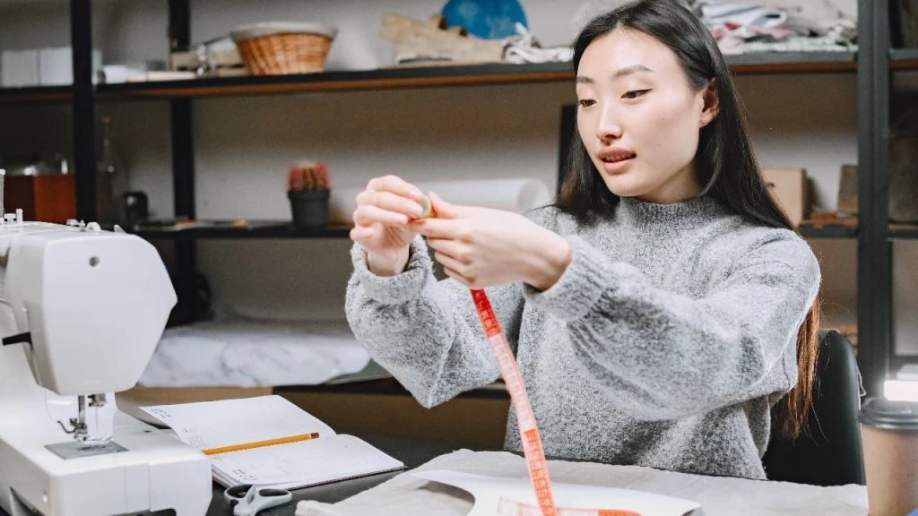 Designer using a measuring tape for sweater measurement chart preparation in a studio.
