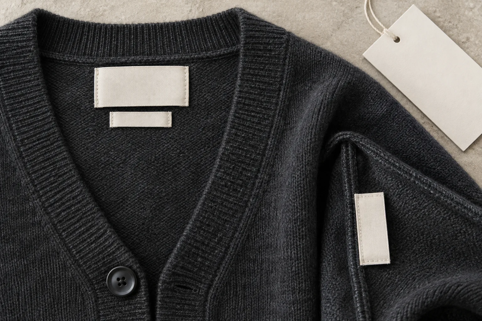 Charcoal cardigan neckline and inside seam showing blank ivory fabric labels beside a blank paper tag
